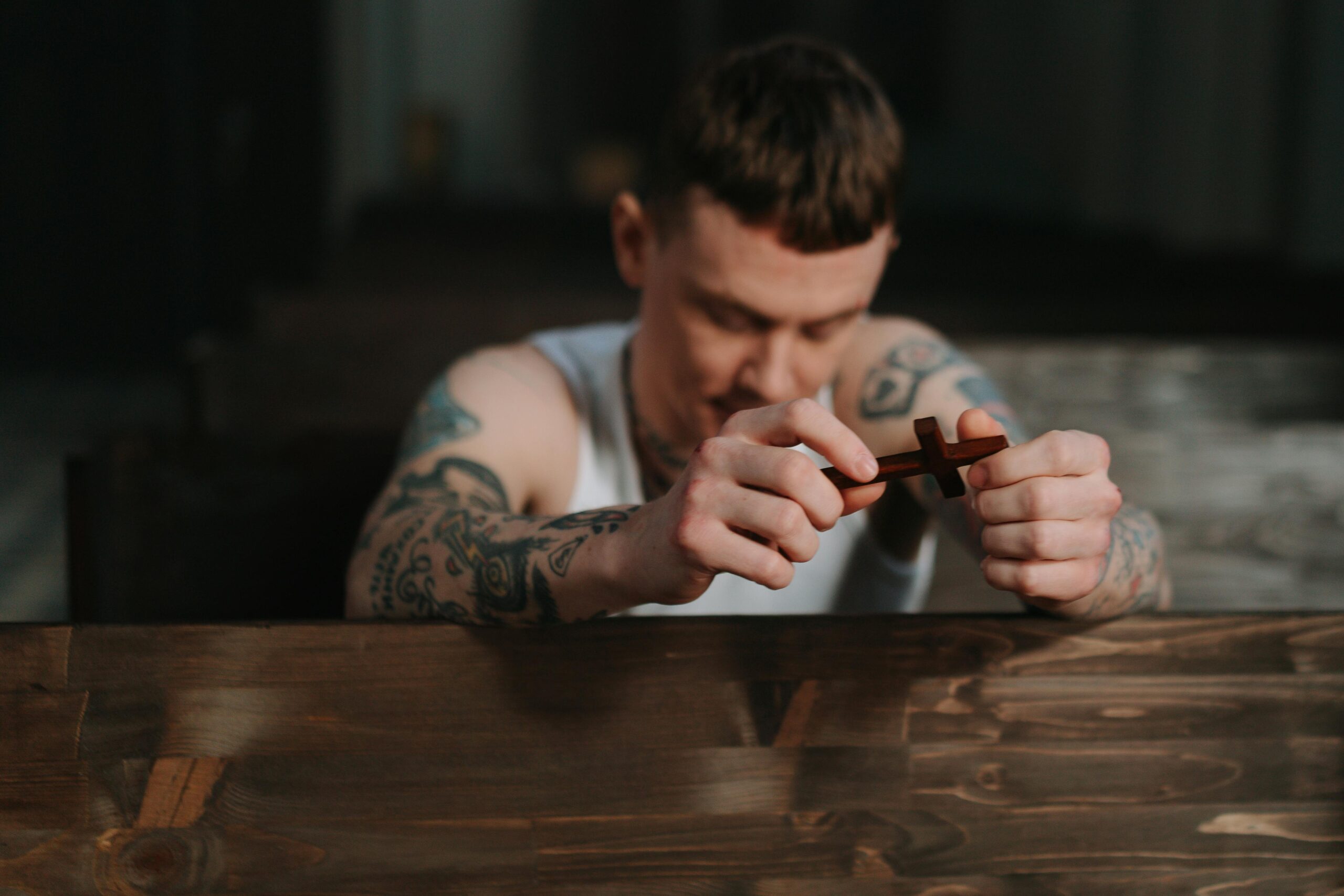 Tattooed man praying with a wooden crucifix inside a dimly lit church, symbolizing faith and reflection.
